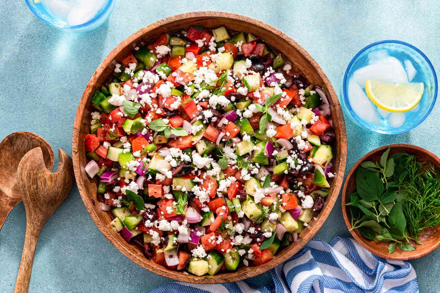 Salade grecque facile dans un bol en bois, et dans les environs, deux verres d'eau citronnée glacée, des ustensiles de service à salade, un bol d'herbes et un torchon de cuisine rayé blanc et bleu