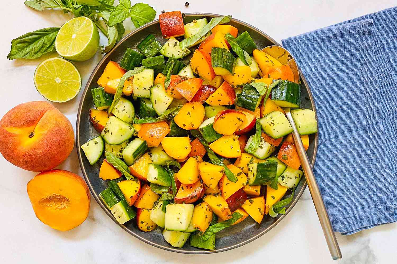 Salade d'été de pêches et de concombres dans un bol avec une cuillère de service. Un linge bleu est à droite et des pêches, des limes et du basilic sont à gauche du bol.