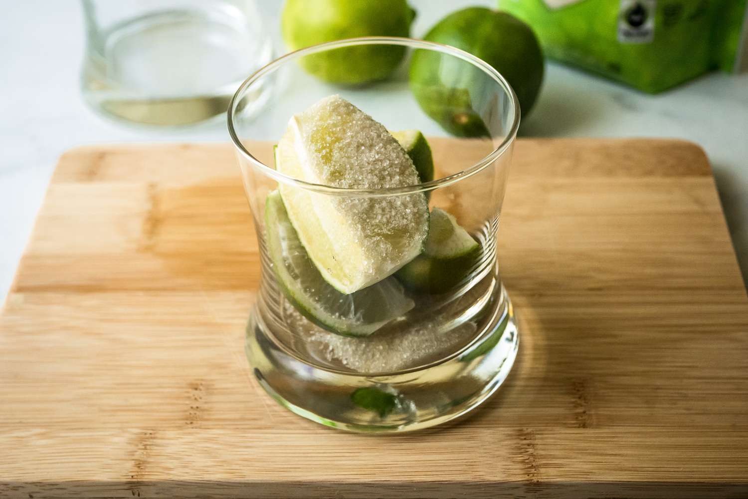 Quartered limes and sugar in a rocks glass set on a cutting board to make a lime caipirinha