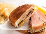 Two halves of a traditional cuban sandwich are resting on parchment paper and a wooden cutting board. Ridged potato chips and a pickle are in the background.