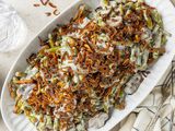 Slow Cooker Green Bean Casserole Topped with Crispy Shallots on a Platter Surrounded by Utensils, a Kitchen Towel, and a Glass of Water 