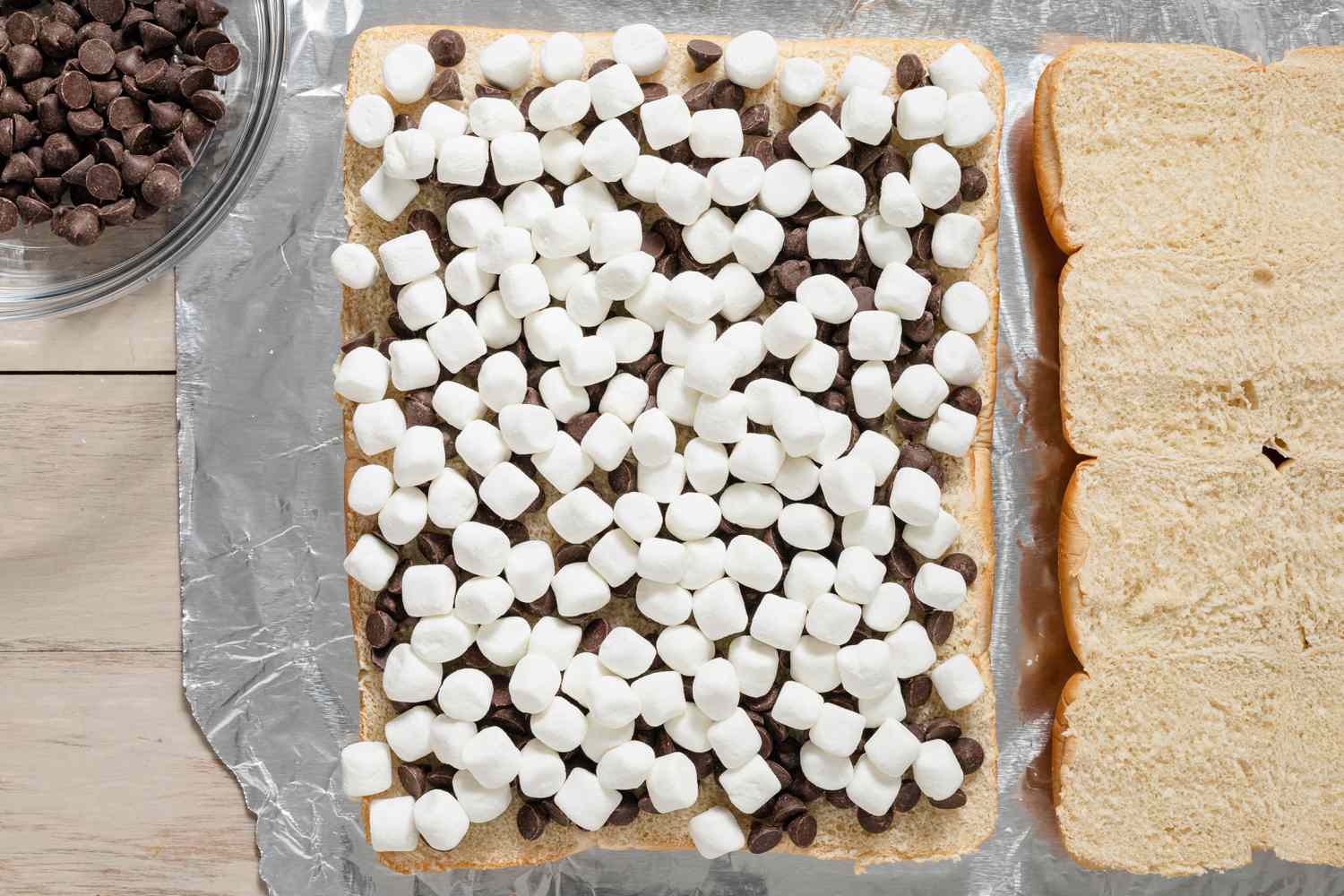 Pépites de chocolat surmontées de mini guimauves sur le même pain à slider coupé. À gauche, un bol avec plus de pépites de chocolat et à droite, l'autre pain coupé pour la recette de sliders s’mores grillés