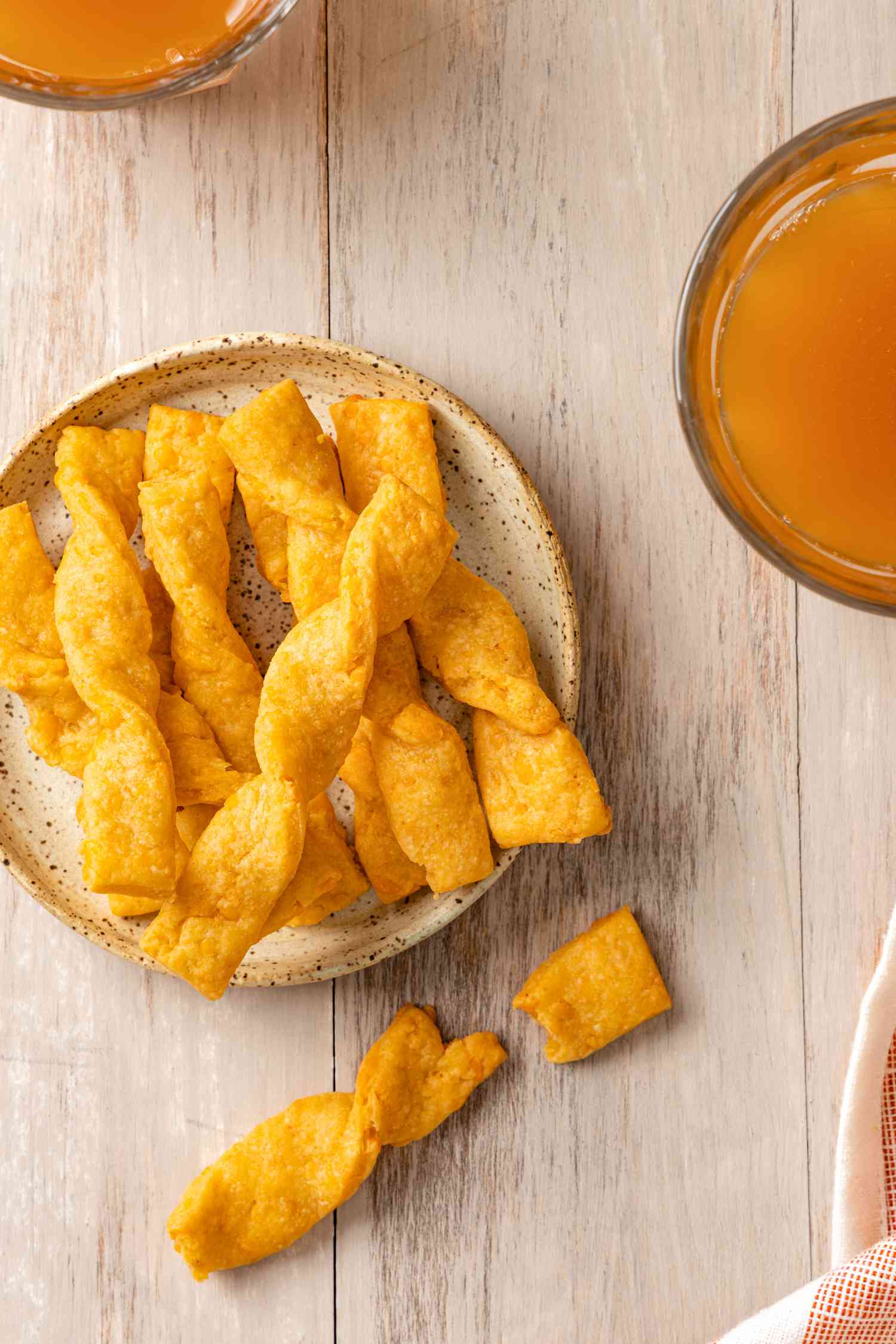 Twisted Cheese Straws on a Plate Next to Two Drinks and a Kitchen Towel. One Twisted Cheese Straw Is Broken in Half Next to the Plate.