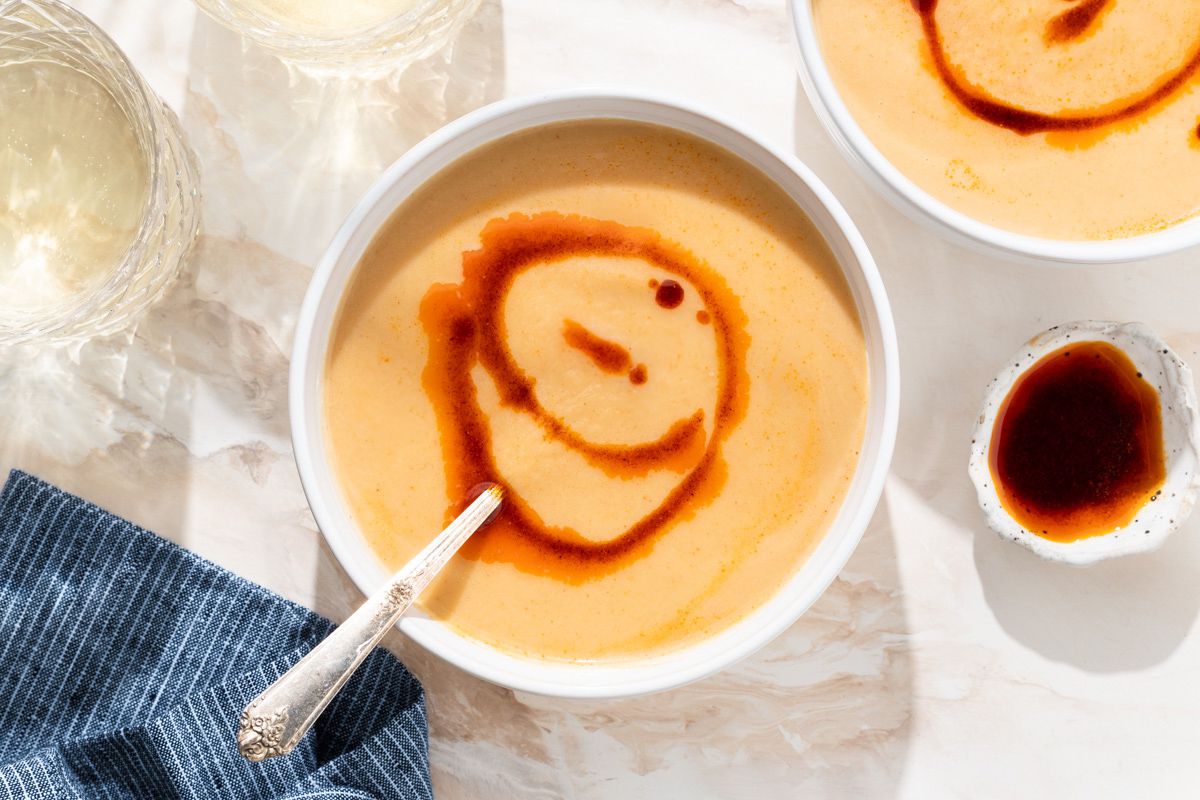 Vue de dessus d'un bol blanc de soupe de chou-fleur arrosée d'huile de paprika à côté d'un petit bol d'huile de paprika, d'une nappe à rayures bleues et de deux verres à boire transparents