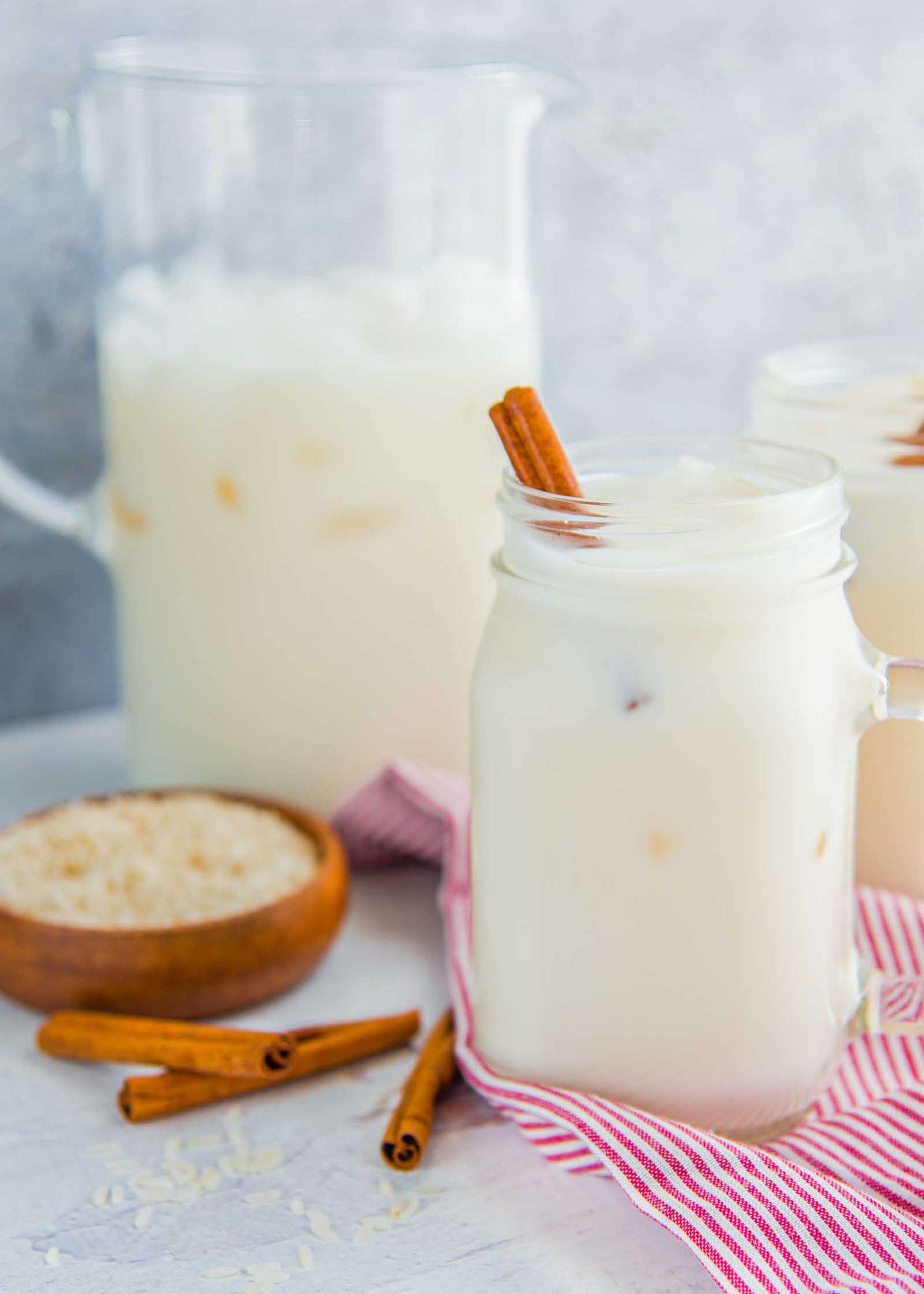 A large mason jar of easy homemade horchata with a cinnamon stick inside is in front of a similar glass and a pitcher of homemade horchata. A small bowl of rice and cinnamon sticks are to the left.