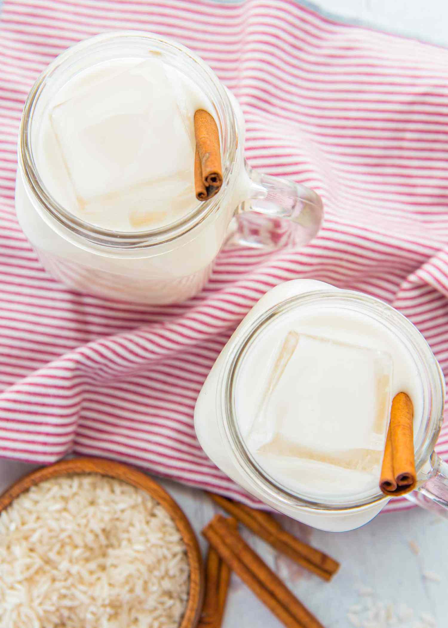 Overhead view of two mason jar mugs of easy homemade horchata over ice with cinnamon sticks inside. A pink striped linen is underneath. Additional cinnamon sticks and a small bowl of rice are in the lower left corner.