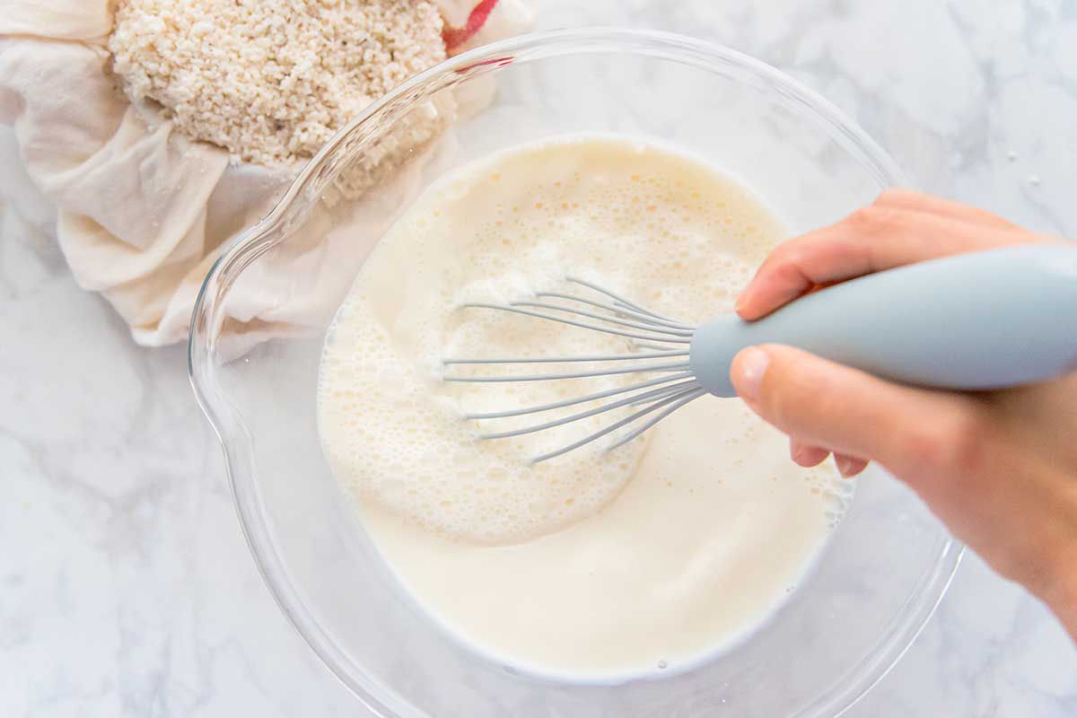 A whisk is combining the ingredients to show how to make horchata. Rice in a cheesecloth is behind the glass bowl of horchata ingredients.