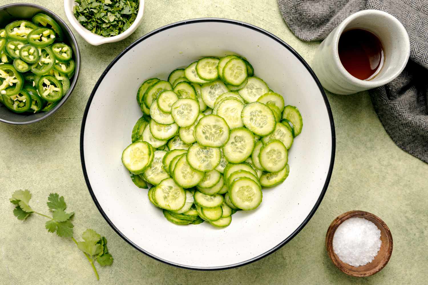 Concombres tranchés dans un bol à côté d'une tasse de sirop d'agave et de petits bols avec des tranches de jalapeño, des herbes et du sel pour la salade épicée de concombre et de jalapeño