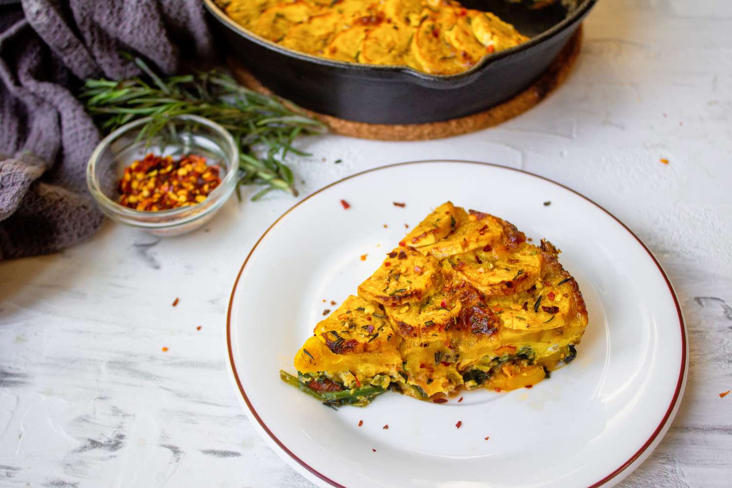 Portion de frittata de banane plantain sur une assiette entourée de romarin, flocons de piment et d'une poêle en fonte