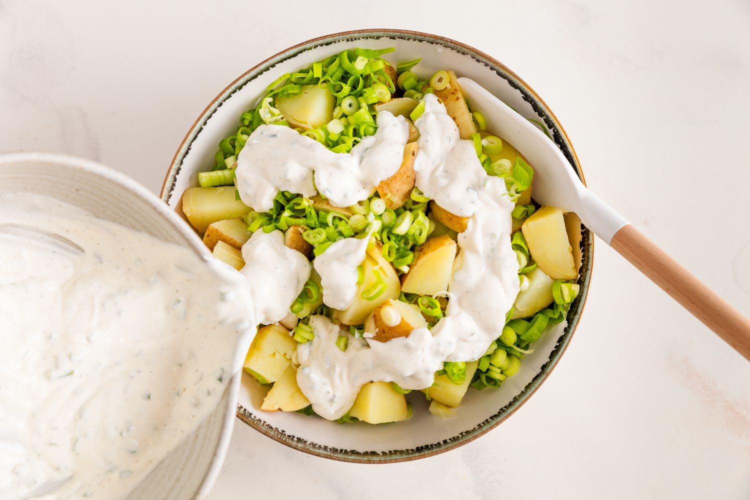 Vinaigrette à la crème aigre et à l'oignon versée sur les pommes de terre et les tranches d'oignons verts dans un bol avec une spatule en caoutchouc