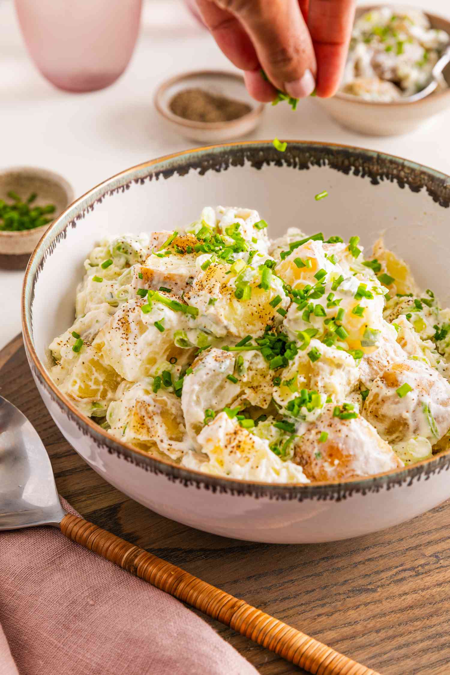 Ciboulette tranchée saupoudrée sur un bol de salade de pommes de terre à la crème aigre et à l'oignon, entouré d'un bol d'assaisonnement, d'un verre d'eau, d'un bol de ciboulette, d'une pile d'assiettes avec des ustensiles, et d'une serviette de table rosée