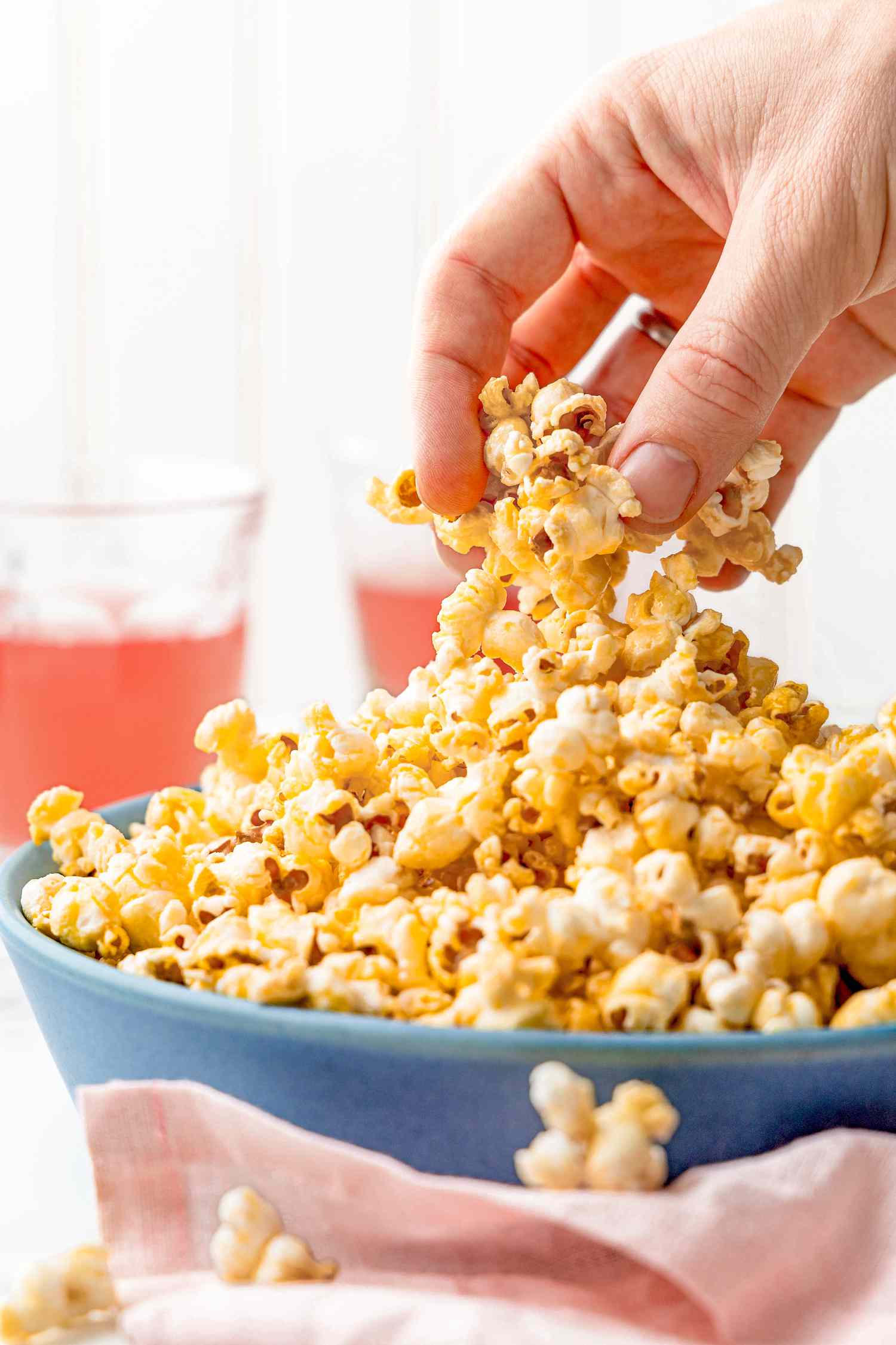 Hand grabbing some of the honey popcorn from a bowl filled with honey popcorn, and in the surroundings, two glasses of pink lemonade and a pink table napkin