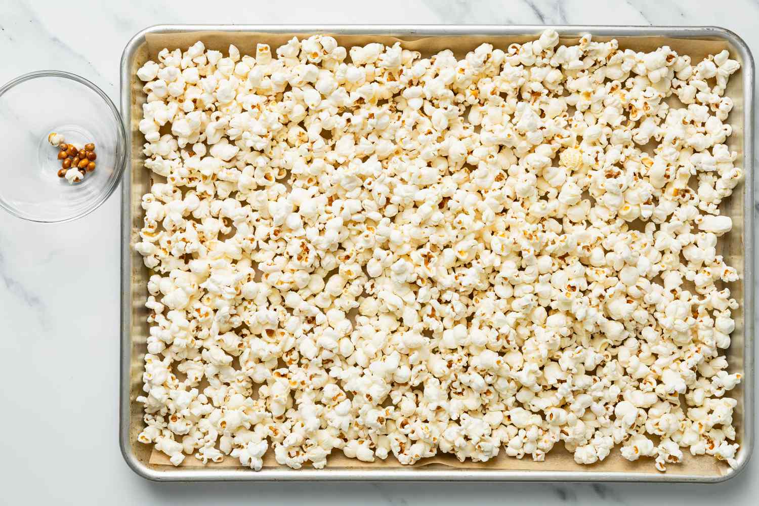 Popcorn on a parchment paper lined tray next to a bowl with unpoped kernels for honey popcorn recipe