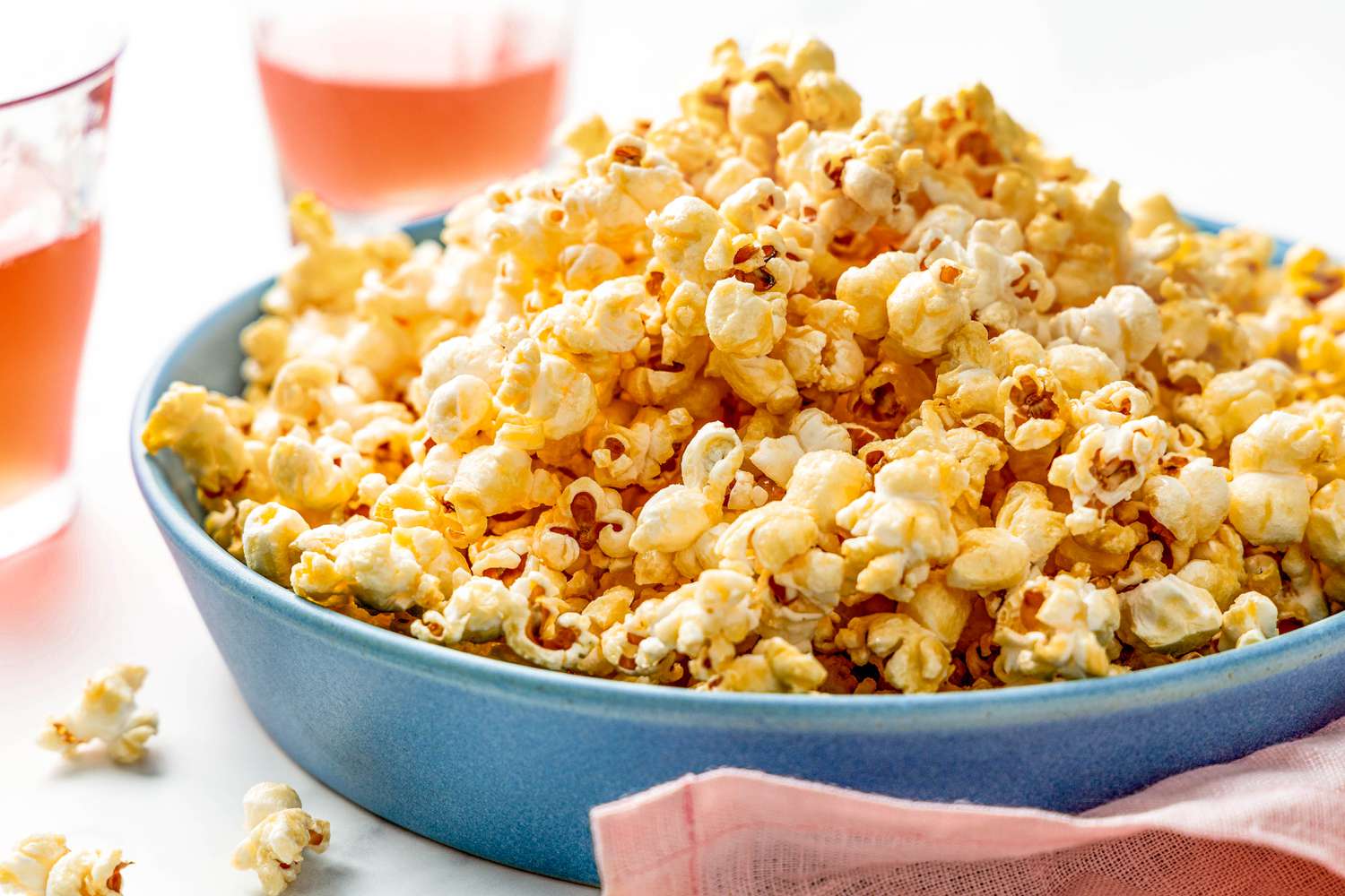 Bowl of honey popcorn surrounded by glasses of pink lemonade, a pink table napkin, and a few pieces of popcorn on the counter