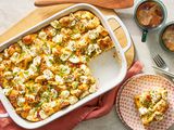 Everything bagel sandwich casserole in a casserole dish at a table setting with a serving on a plate, utensils, and cups of coffee