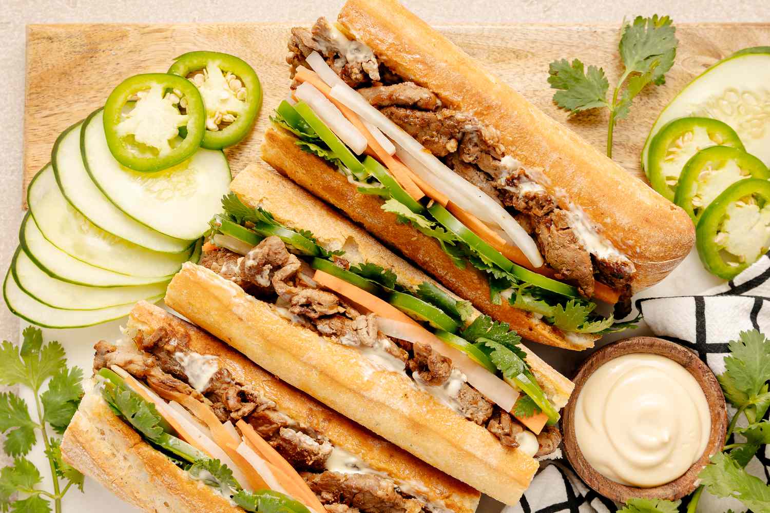 Easy Pork Bánh Mì Surrounded by Slices of Cucumbers and Jalapenos and a Bowl of Mayo