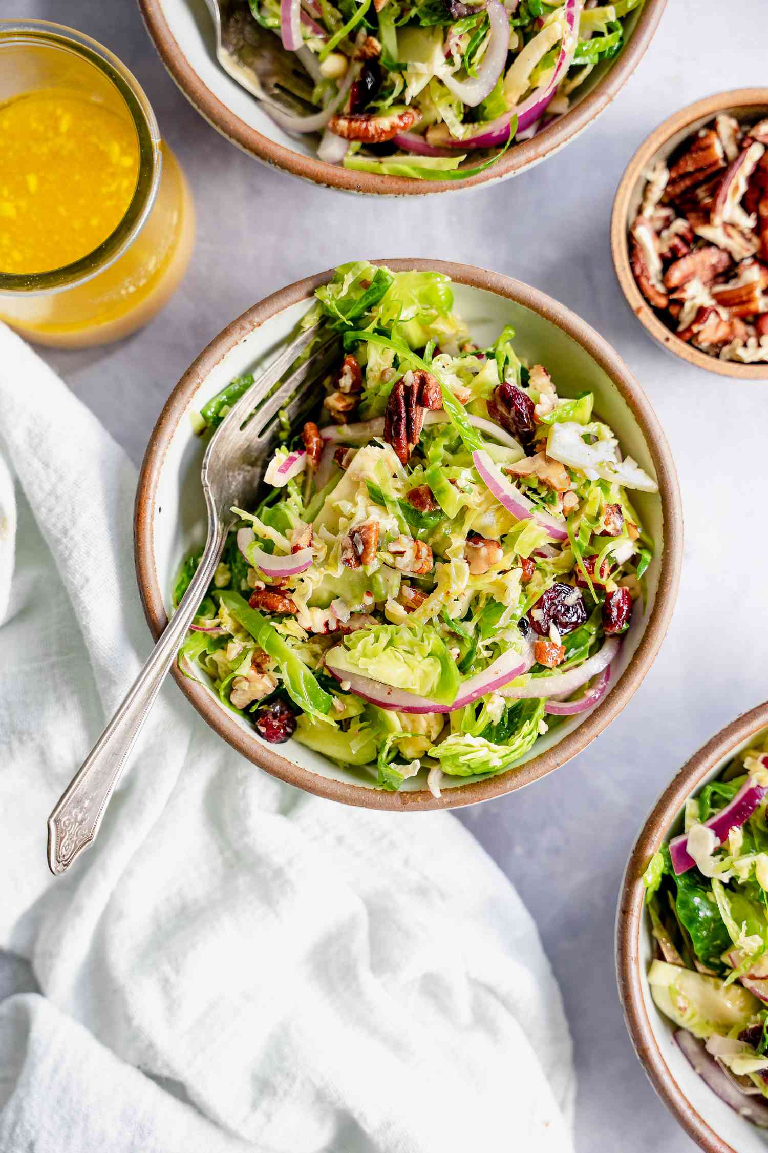 Brussel Sprout Cole Slaw in Three Bowls Next to a Jar of Dressing and Bowl of Pecans