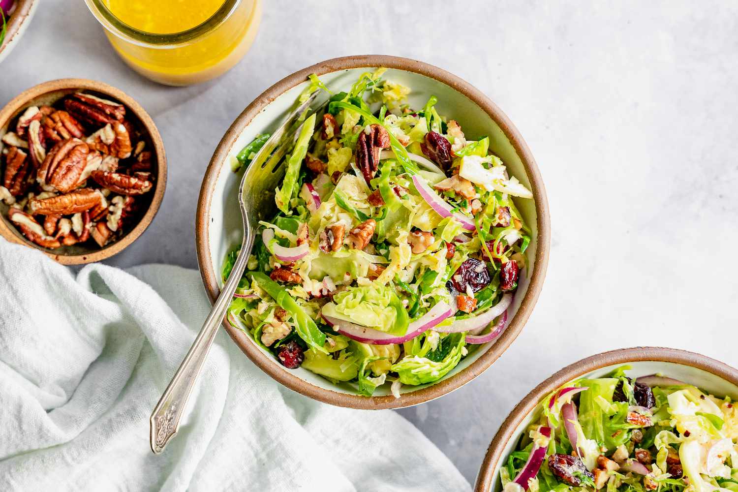 Brussel Sprout Cole Slaw in Three Bowls Next to a Jar of Dressing and Bowl of Pecans