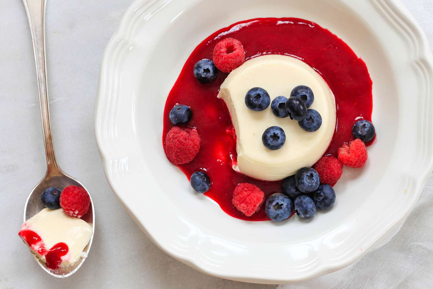 Panna Cotta garnie de baies d'été dans un bol et sur le comptoir une cuillère de panna cotta avec une framboise et une myrtille