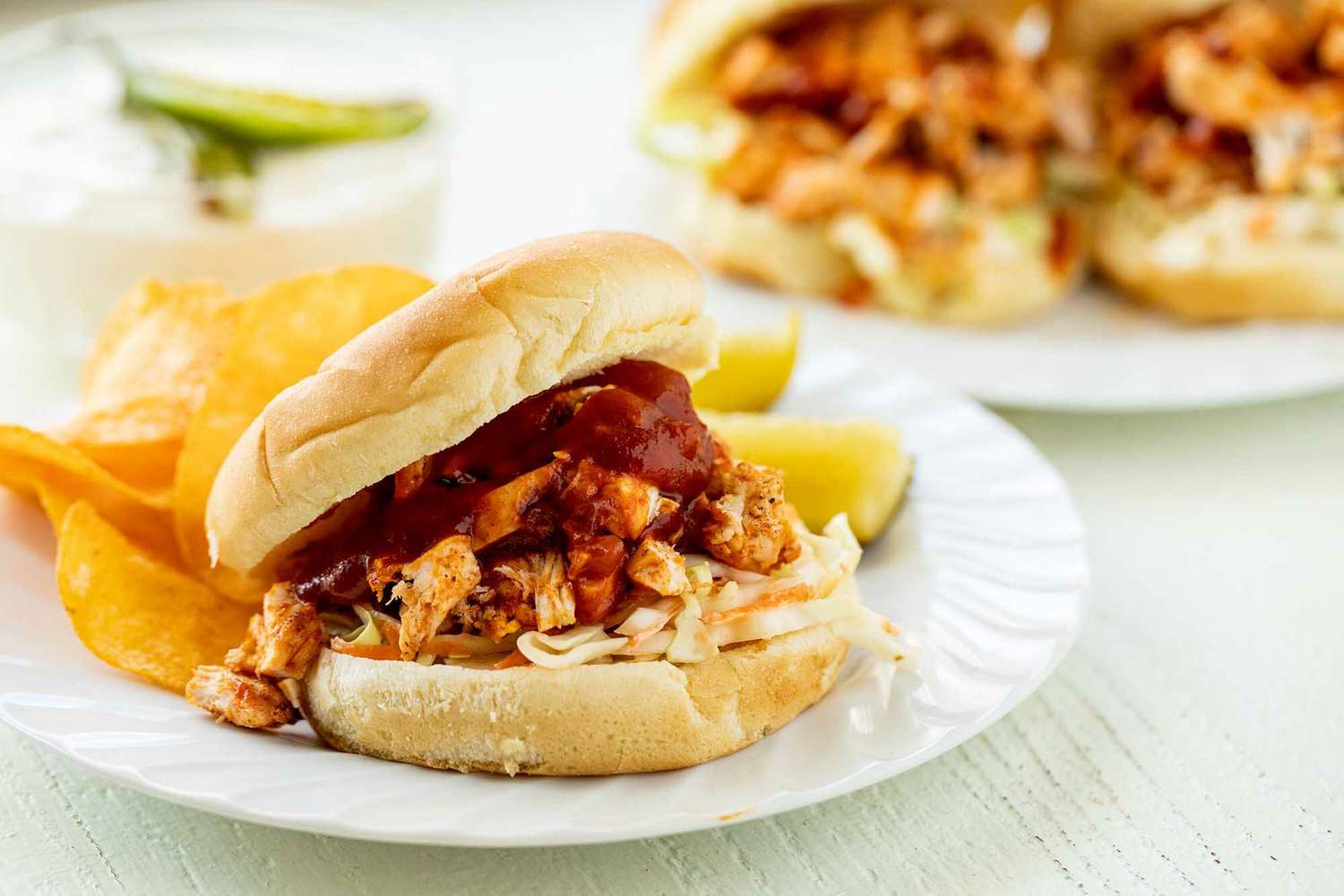 Une Assiette avec des Cornichons, des Chips et un Sandwich au Poulet BBQ Effiloché avec Chou Marinée et Garni de Sauce BBQ. Une Deuxième Assiette avec Deux Sandwichs et de la Mayonnaise de Serrano se Trouve Derrière.