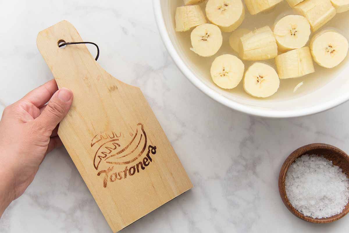 Des plantains coupés trempant dans un bol d'eau avec une tostonera à proximité.