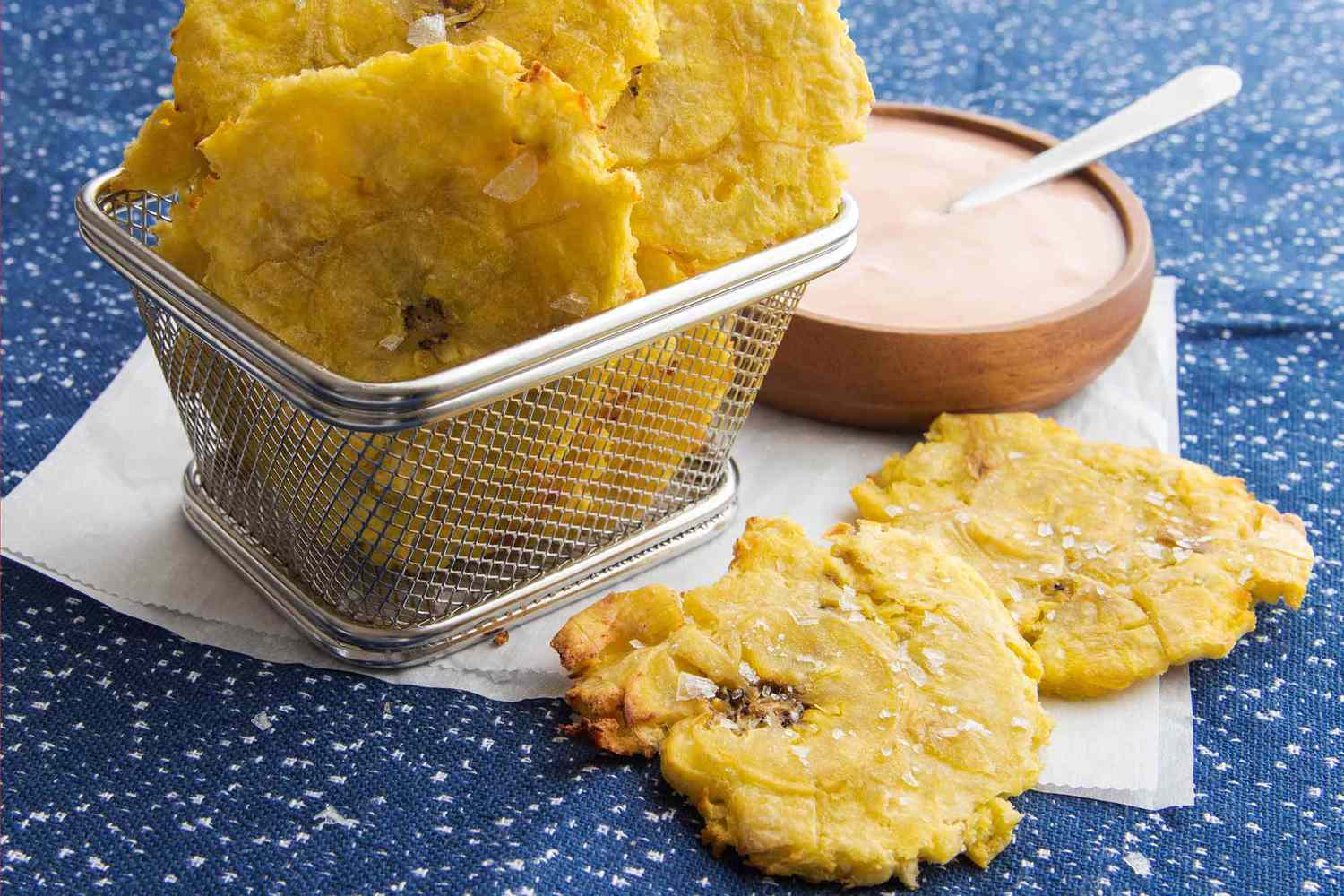 Tostones au four à air dans un panier posé sur du papier sulfurisé avec un bol de sauce à côté.