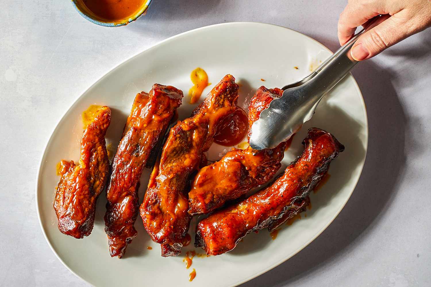 A hand holding tongs grabbing a country-style rib of a serving plate with more ribs