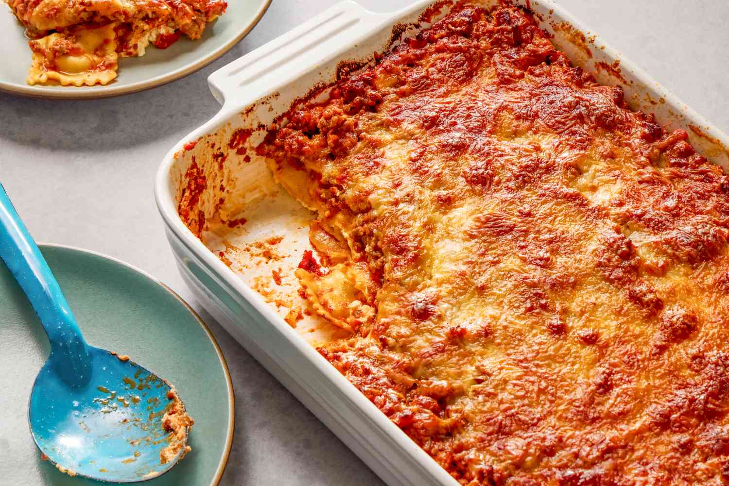 Vue en angle d'un plat à gratin blanc de casserole de ravioli à un million de dollars avec une portion retirée et sur une petite assiette à côté d'une cuillère de service sur une petite assiette.