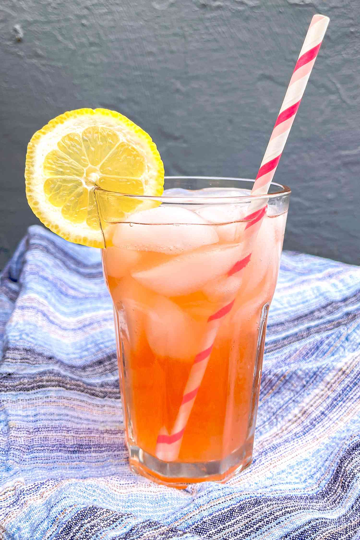 Glass of Strawberry Lemonade With a Lemon Slice on the Glass Rim and a Straw Sitting on a Blue Kitchen Towel