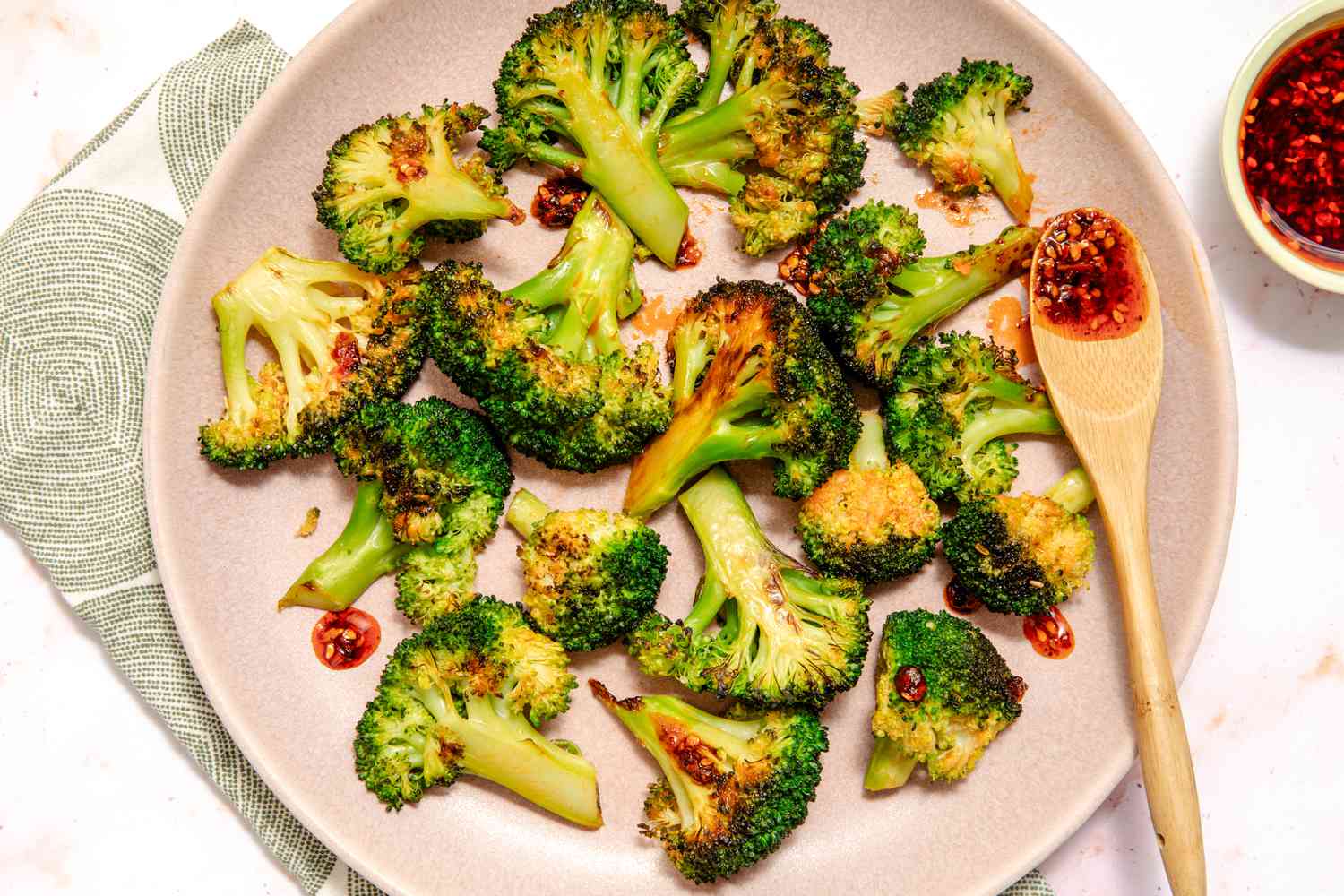 Vue de dessus d'une assiette rose de fleurettes de brocoli recouvertes de chili croustillant, avec une petite cuillère en bois avec du chili croustillant, le tout reposant sur une serviette en tissu et un comptoir en marbre