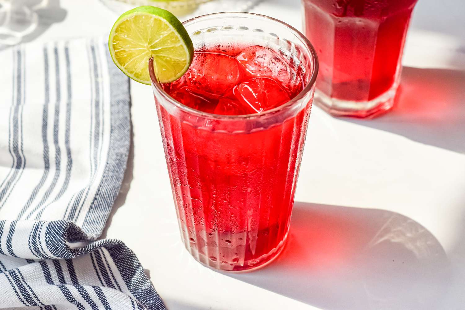 Agua de Jamaica (thé d'hibiscus) servi avec de la glace et une tranche de lime à côté d'une serviette de cuisine et un autre verre de thé d'hibiscus