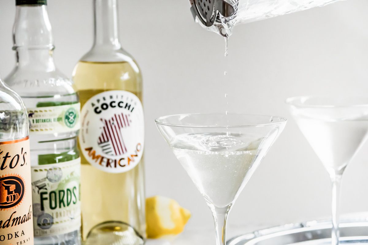 Cocktail Vesper versé dans un verre à martini depuis un verre à mélange et à gauche, des bouteilles de vodka, de gin et d'Americano