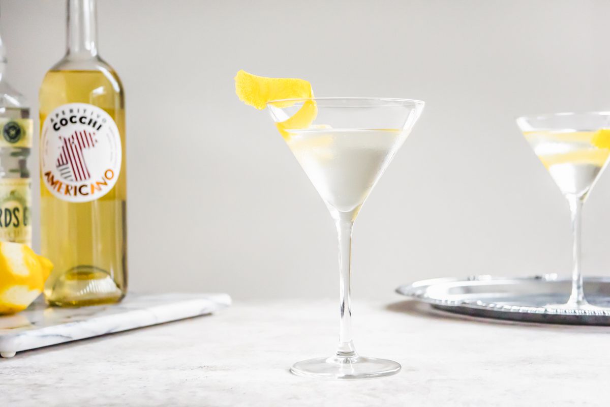 Cocktail Vesper sur le comptoir et en arrière-plan, à gauche, un plateau en marbre avec des bouteilles d'Americano et de gin et à droite, un autre verre sur un plateau en argent