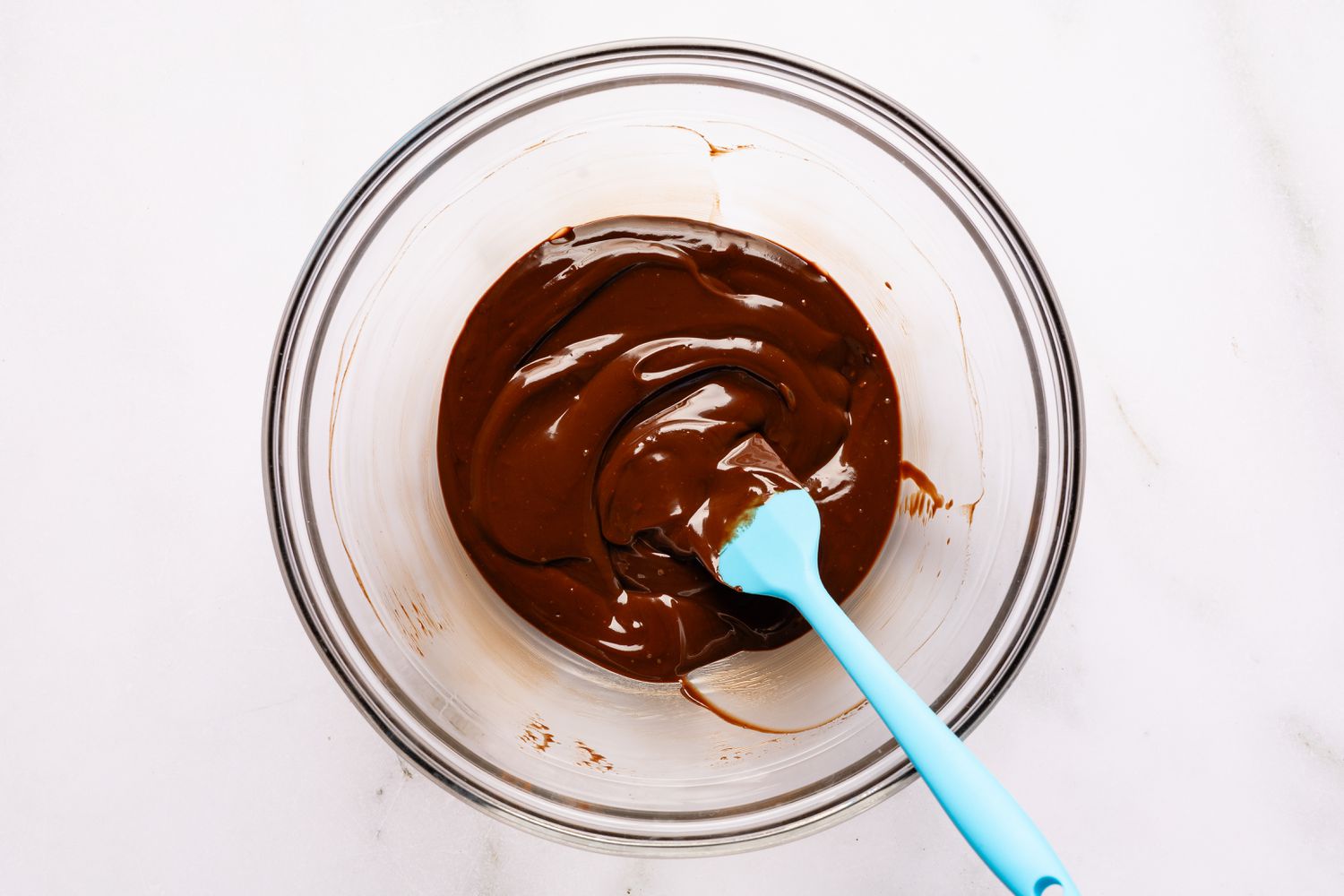 Vue de dessus d'un bol en verre transparent avec le mélange de crème et de chocolat fondu ainsi qu'une spatule bleue pour la recette de gâteau Brooklyn Blackout facile