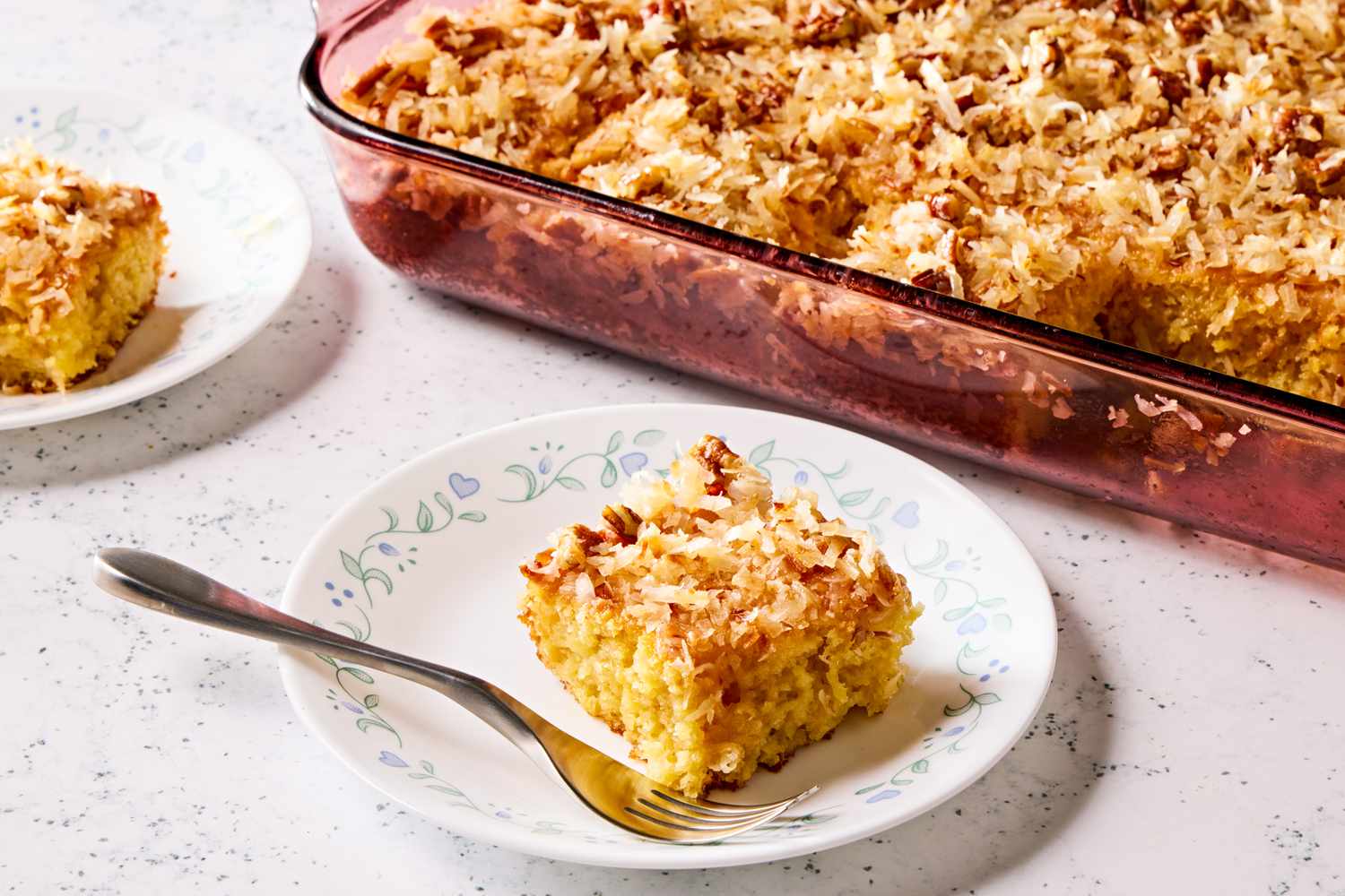 Vue en angle d'un plat de cuisson en verre contenant du gâteau à l'ananas et à la noix de coco à côté de deux petites assiettes avec des parts