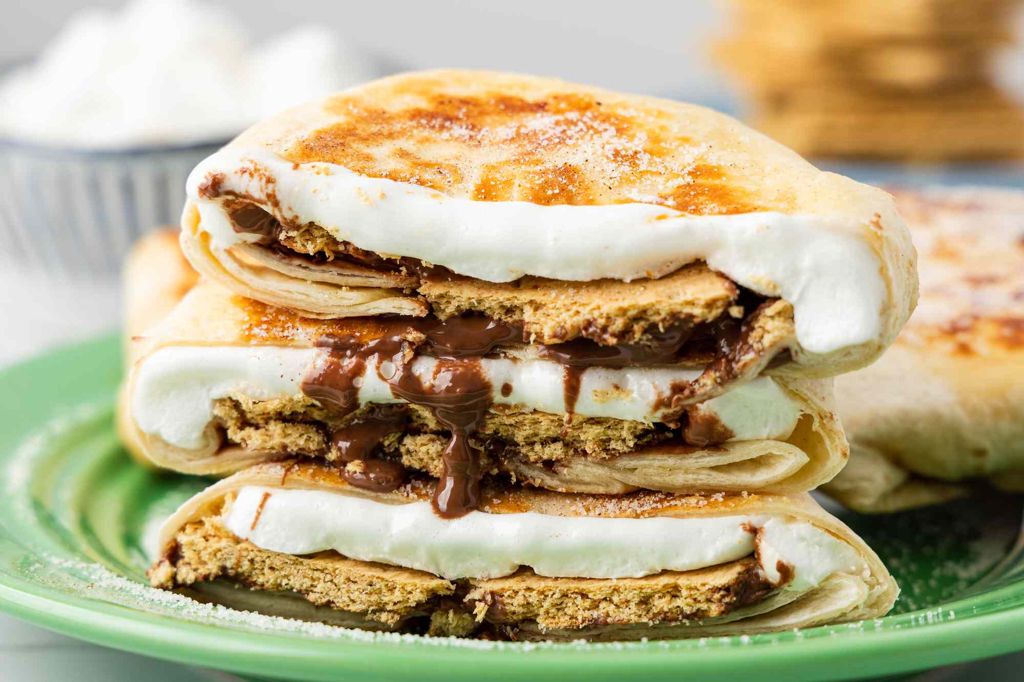 Cross-Section Cut of a S'mores Crunchwrap on a Plate, and in the Background, a Bowl of Mini Marshmallows and a Plate With a Stack of Graham Crackers