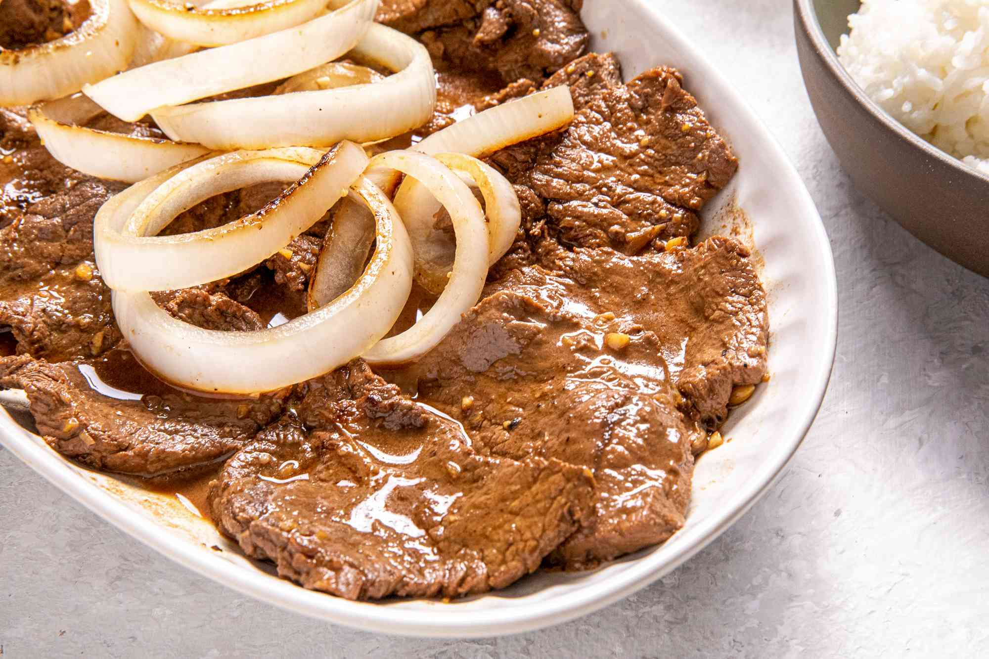 Plate of Bistek Topped With Onions Next to a Bowl of Rice