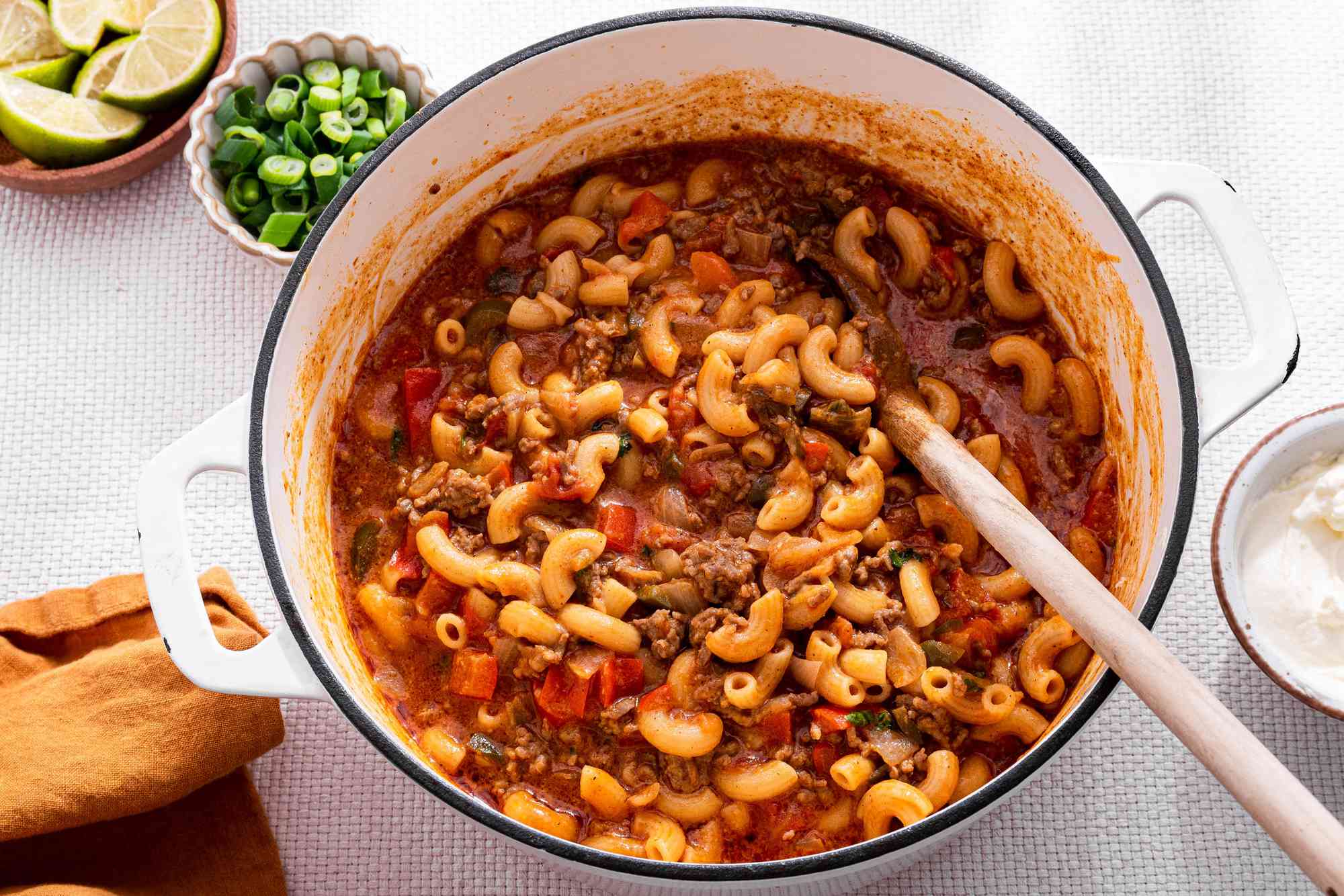 One-Pot Taco Pasta in a Pot, and in the Surroundings, a Bowl With Lime Wedges, a Bowl With Sliced Green Onions, a Bowl With Sour Cream, and a Mustard Colored Table Napkin