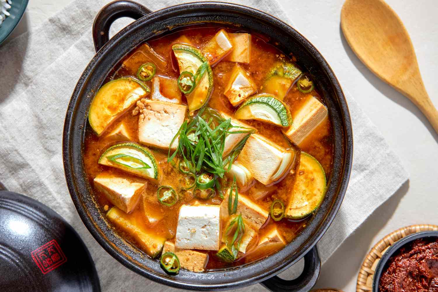 Vue aérienne du Doenjang Jjigae dans une casserole en terre cuite sur un torchon de cuisine blanc, entouré d'un bol de riz, d'un petit bol de sauce et d'une cuillère en bois