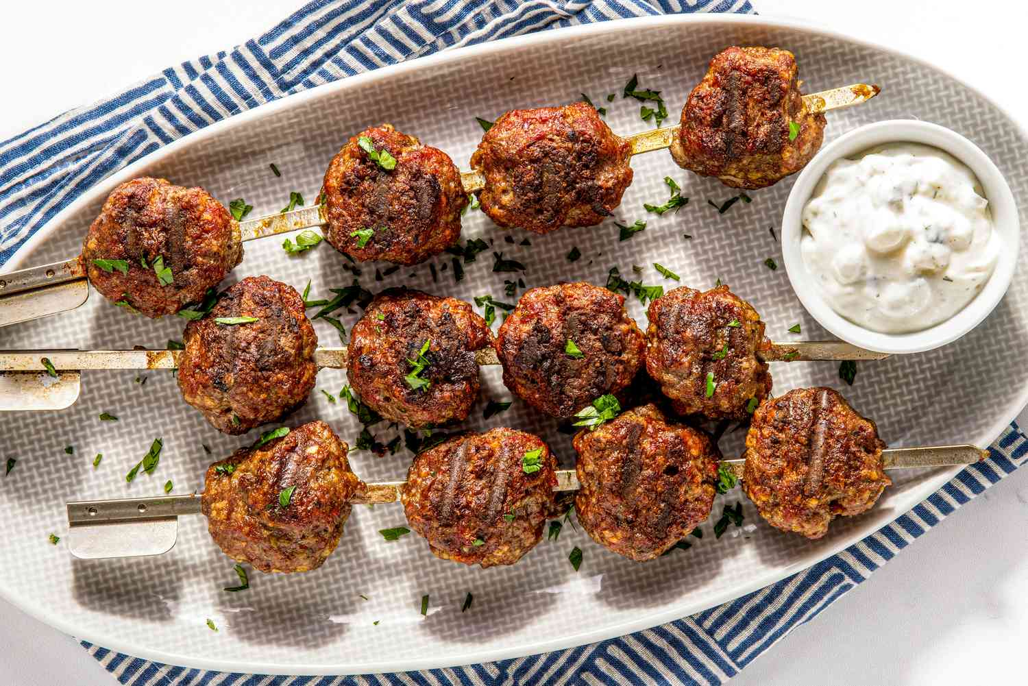 Plat de Boulettes de Viande Grillées Avec un Petit Bol de Tzatziki, Le Tout Sur une Serviette de Cuisine Rayée Bleue et Blanche