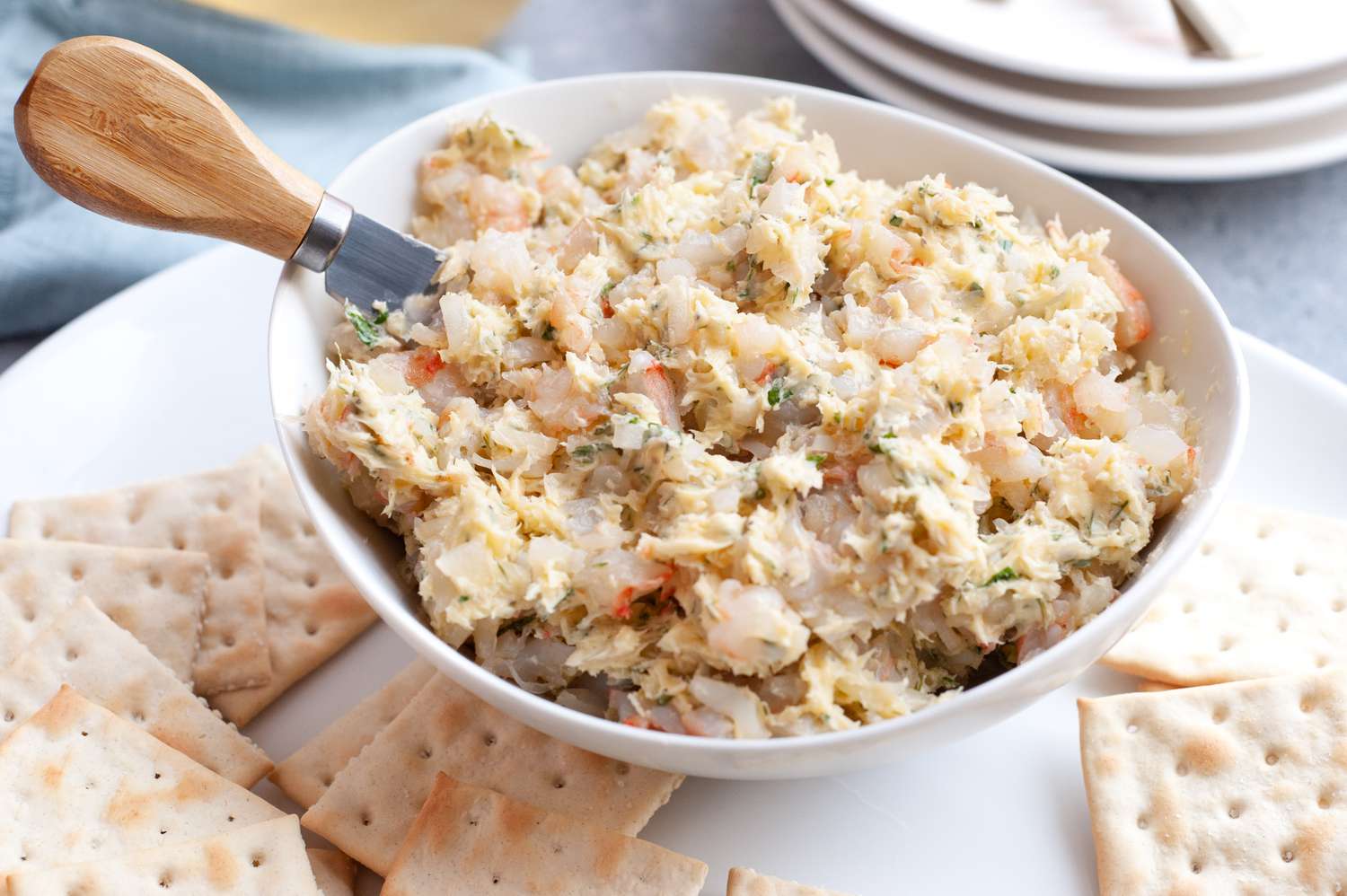 Bowl of Shrimp Butter with a Cheese Knife, Sitting on a Platter of Crackers at a Table Setting with a Glass of Wine, Stack of Plates, and a Kitchen Linen