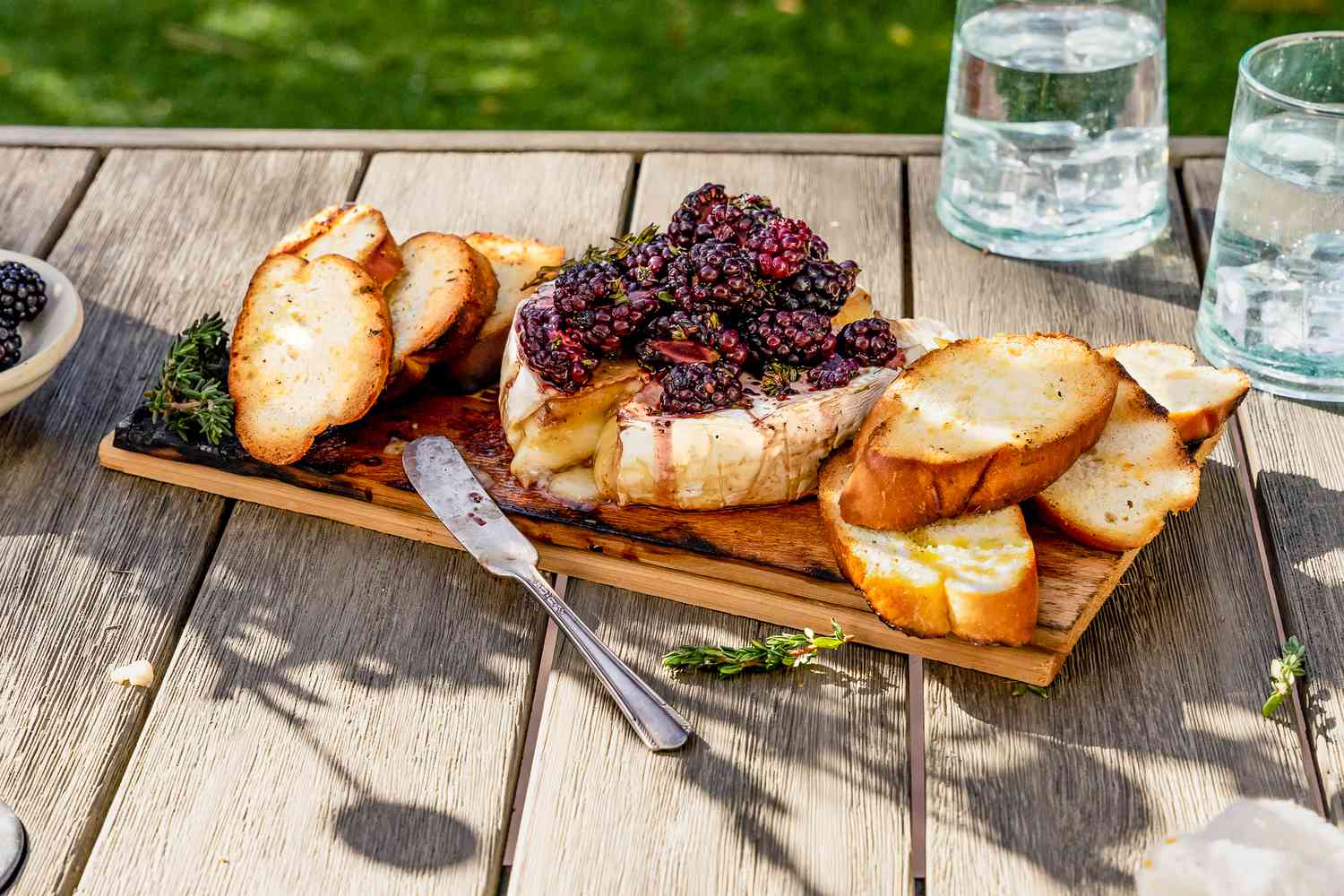 Brie grillé sur planche de cèdre avec mûres et brins de thym, sur le plateau avec crostini, et à côté des verres d'eau