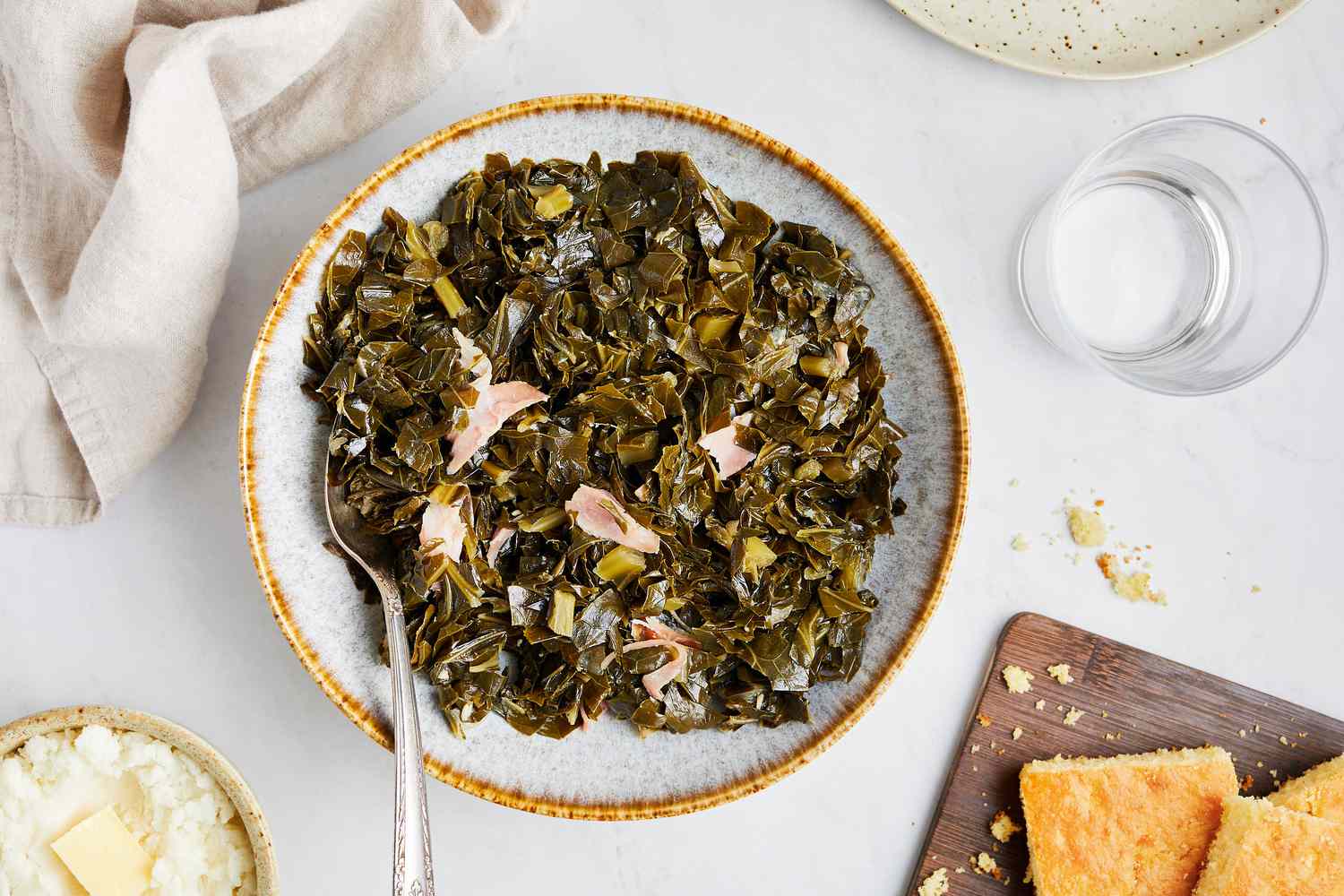 Instant Pot Collard Greens on a Plate with Serving Utensil Surrounded by a Bowl of Mashed Potatoes, Cornbread on a Wooden Tray, and a Glass