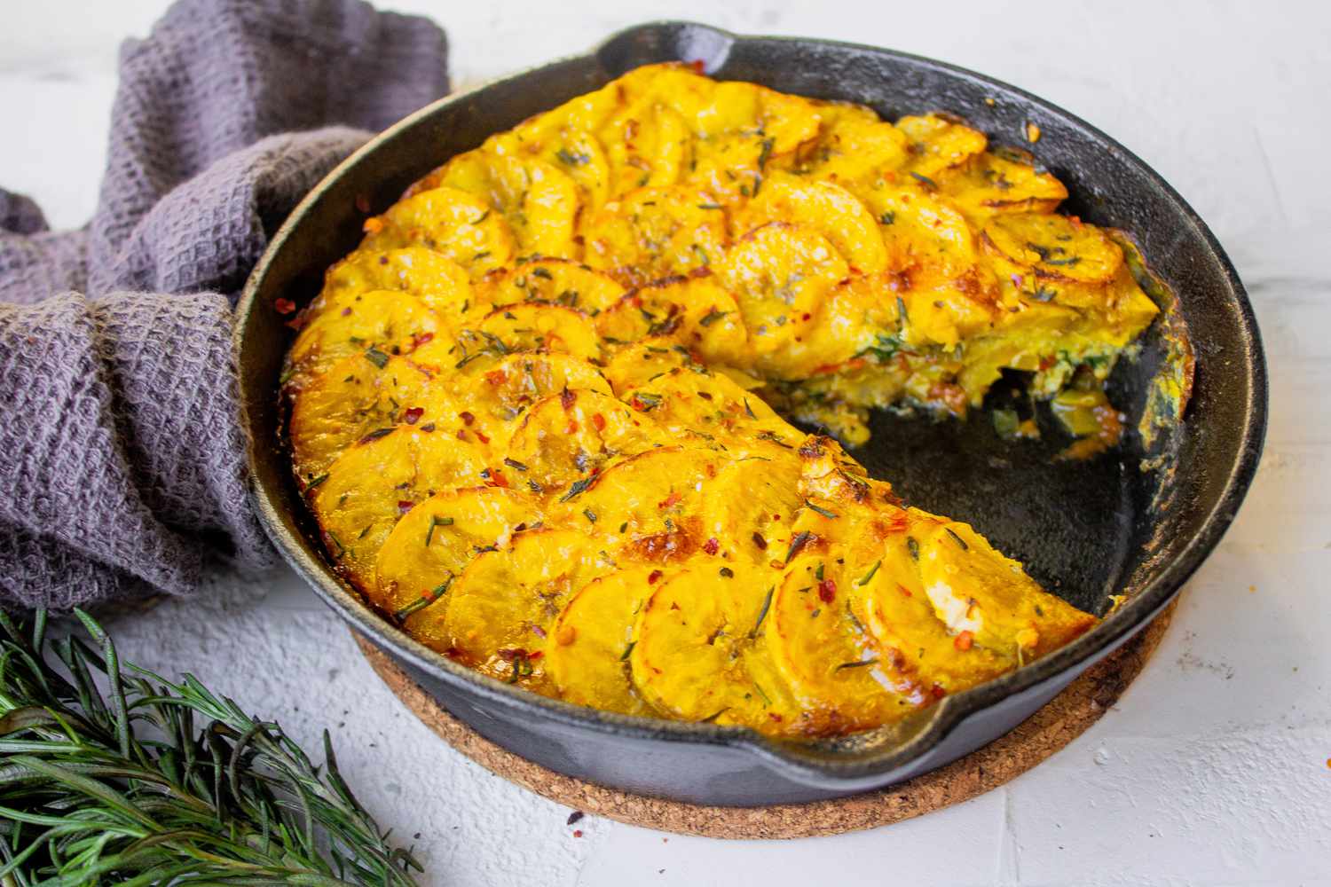 Frittata de banane plantain avec une portion coupée dans une poêle en fonte entourée de romarin et d'une serviette de cuisine