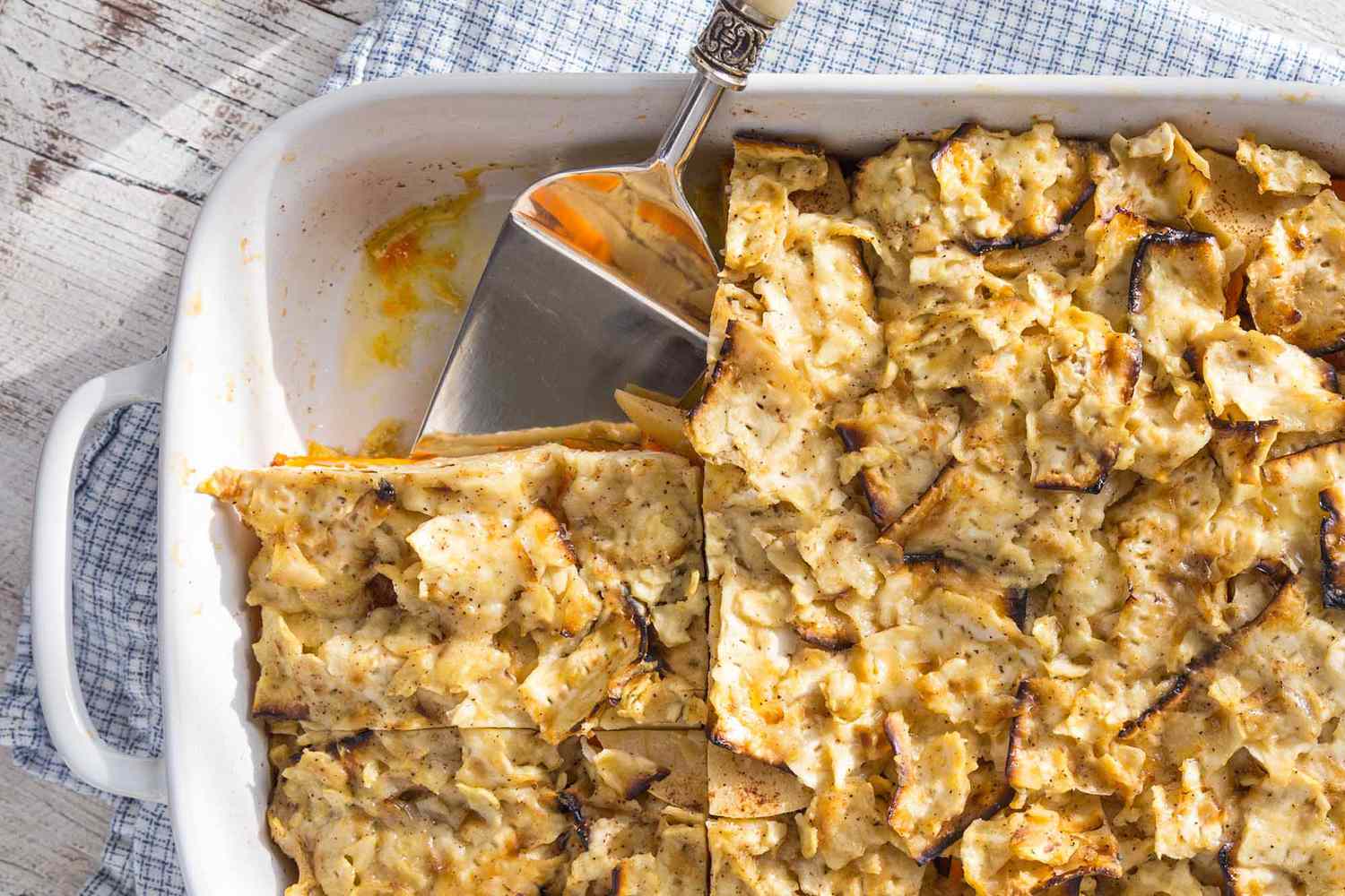 Passover Side Dish with Matzo - casserole dish filled with layers of sweet potatoes and apples, with matzo crackers on top. A slice is missing and a serving spatula is in its place