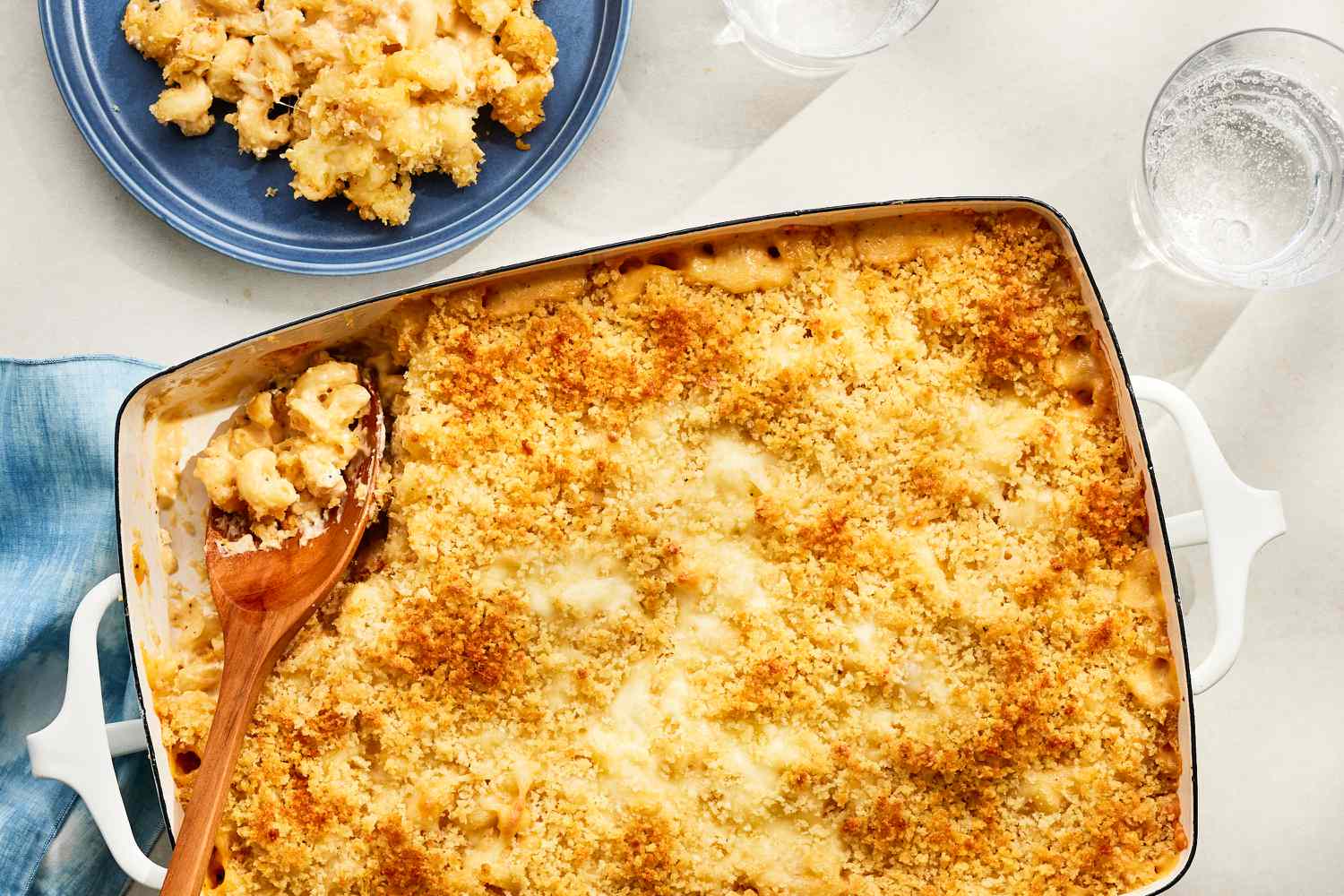 Vue aérienne de macaronis et fromage d'un million de dollars surmontés de panko dans un plat de cuisson avec une cuillère de service dans le coin, le tout à côté d'une petite assiette bleue avec une portion