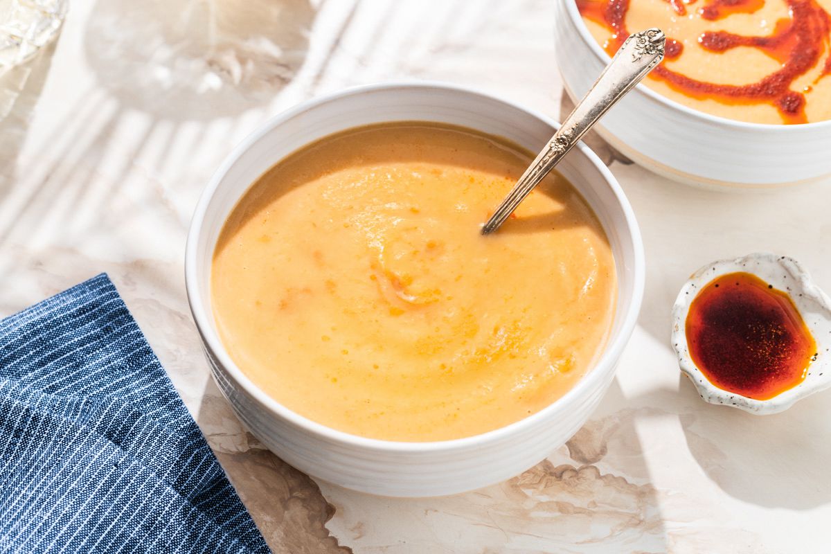 Vue d'angle d'un bol blanc de soupe de chou-fleur à côté d'un petit bol d'huile de paprika, d'une nappe à rayures bleu et de deux verres à boire transparents