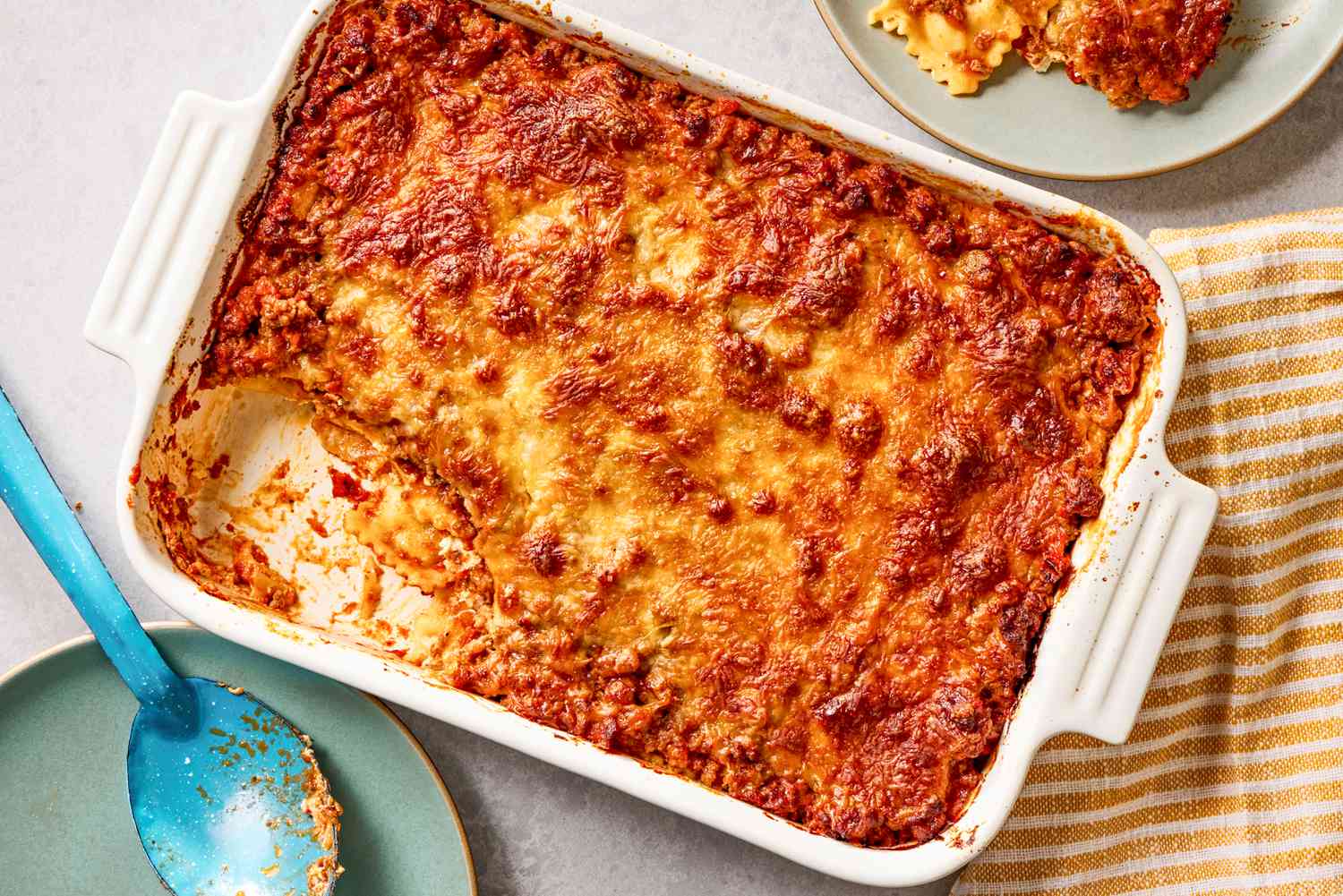 Aperçu aérien d'un plat à gratin blanc de casserole de ravioli à un million de dollars avec une portion retirée et sur une petite assiette, une cuillère de service et une serviette rayée