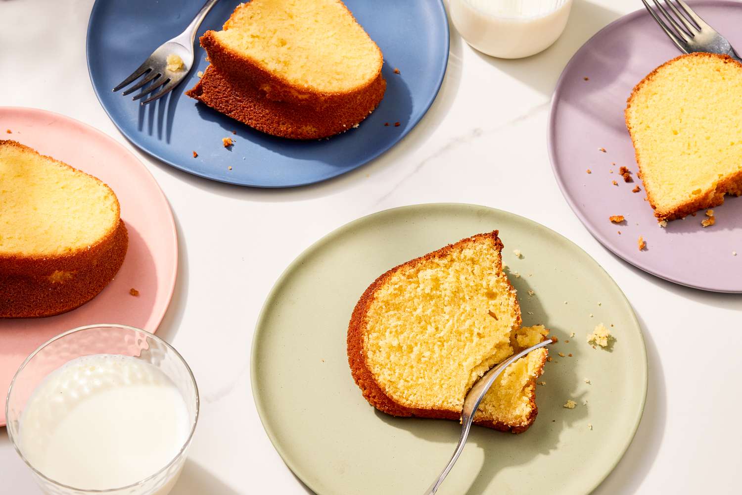 Vue d'ensemble de 4 assiettes avec des tranches de gâteau pound cake et des fourchettes à côté de verres de lait sur un plan de travail en marbre