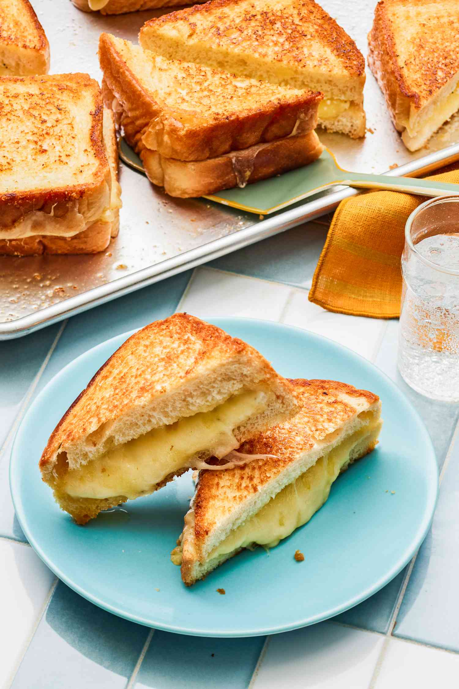 Sheet Pan Grilled Cheese Sandwich Cut in Half and on a Light Blue Plate Next to a Table Napkin and a Sheet Pan With More Grilled Cheese Sandwiches